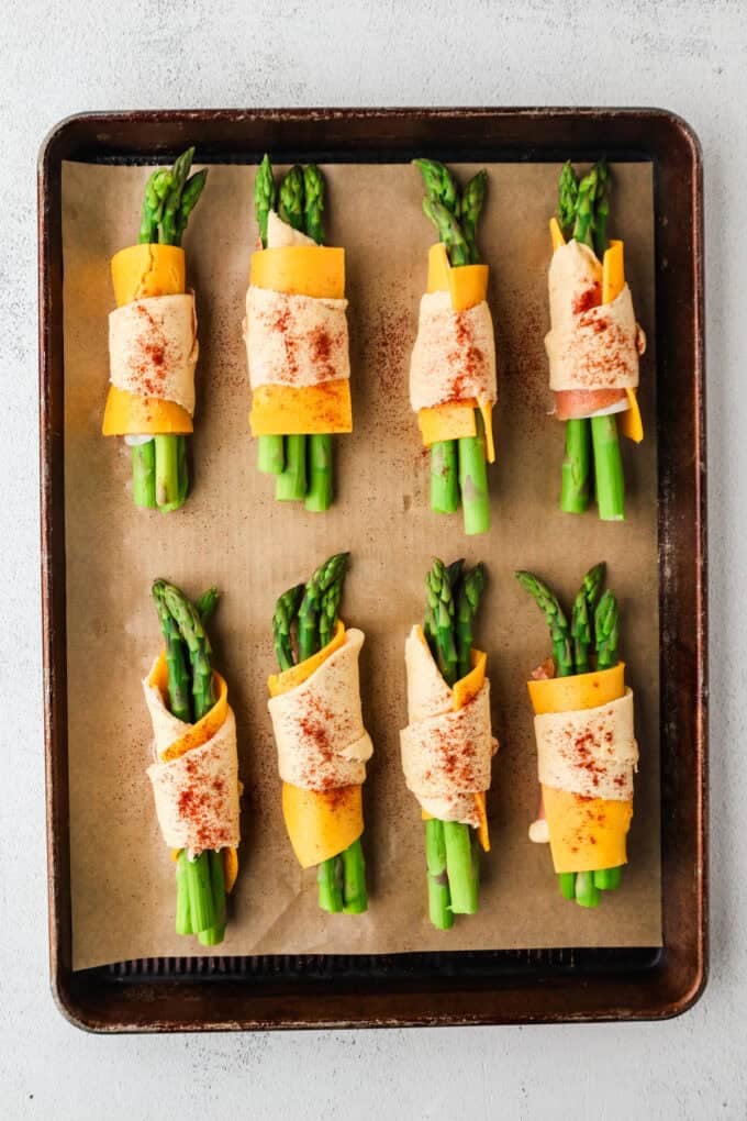 A parchment paper lined baking sheet with two rows of four each asparagus rolls sprinkled with paprika before being baked.