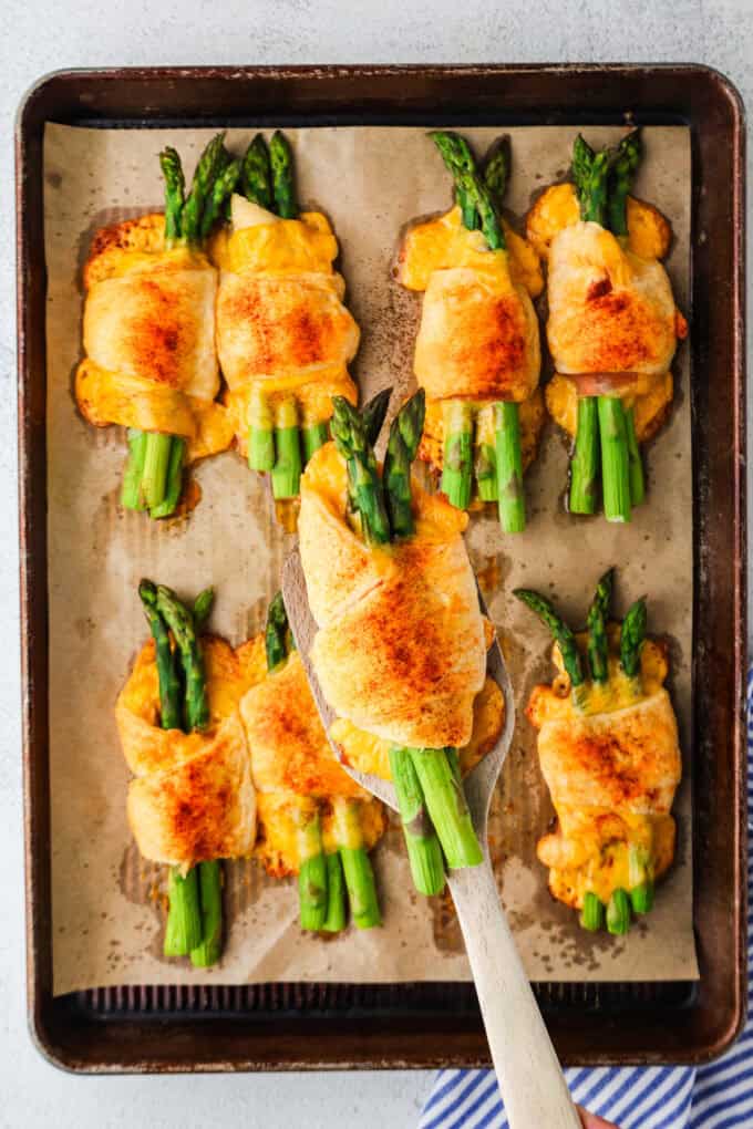 A spatula lifting up a baked asparagus roll from a sheet pan full of rolls.
