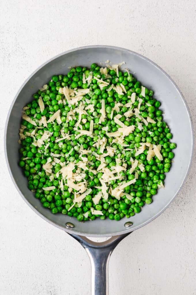 A skillet full of green peas with shallots and shaved Parmesan cheese on top.