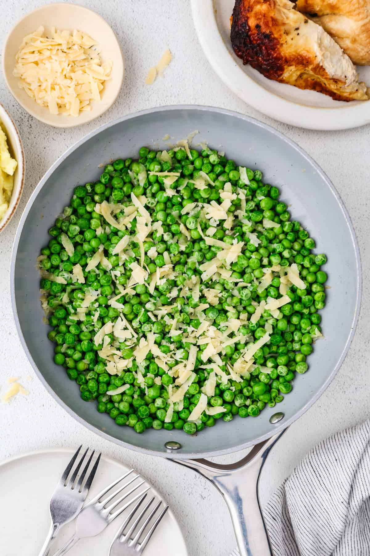 A skillet of green peas with shallots and shaved Parmesan cheese on top with extra Parmesan and a plate of roasted chicken nearby.