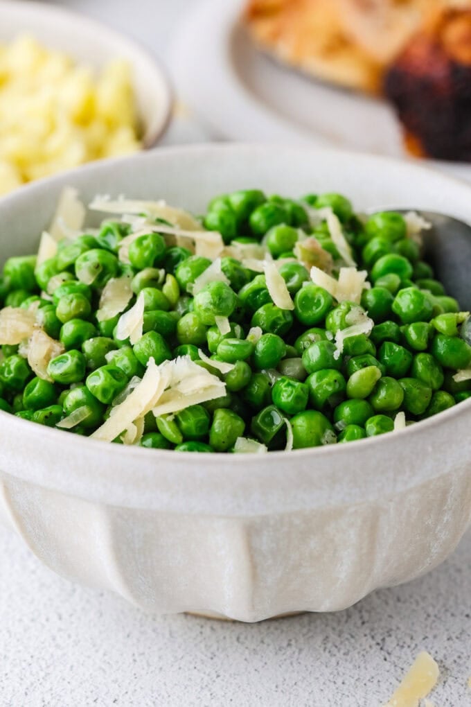 A serving bowl full of green peas with Parmesan cheese and a spoon resting in the side.