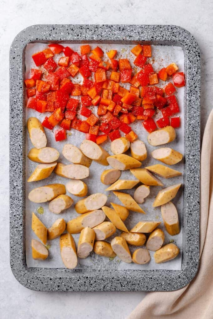 A sheet pan with diced red bell pepper and sliced chicken sausages before being roasted.