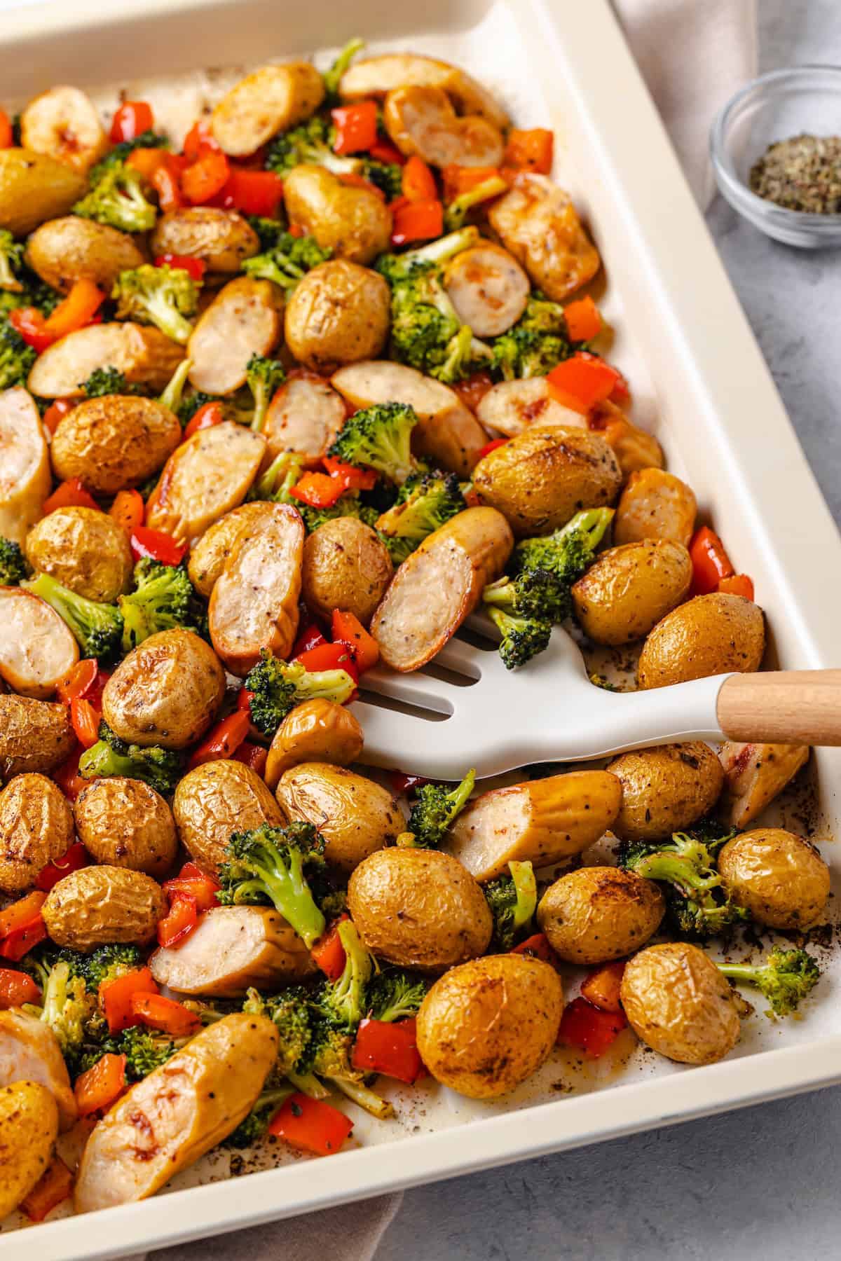 A spatula scooping up chicken sausage and veggies from a sheet pan.