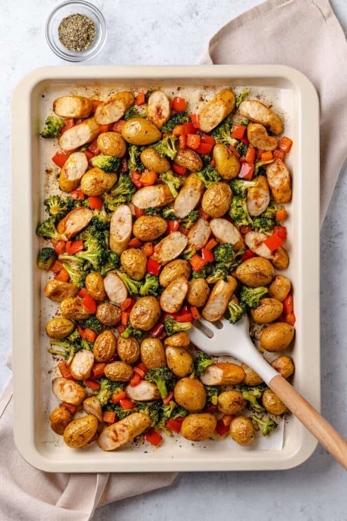A spatula resting on a sheet pan full of potatoes, broccoli and sliced chicken sausage.