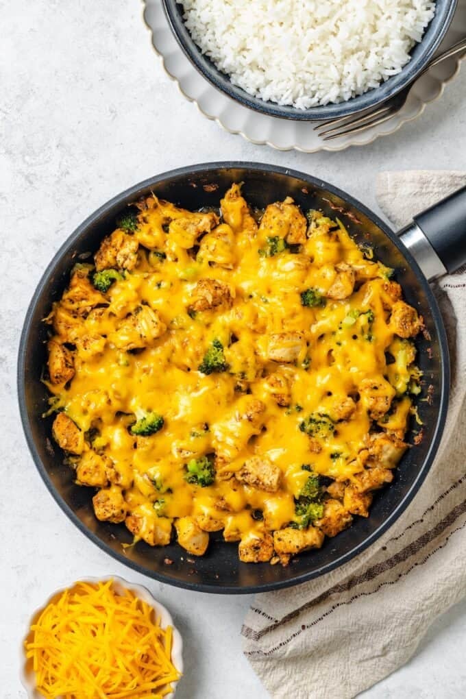 Melted cheddar cheese on top of a chicken and broccoli mixture in a large skillet with a bowl of rice to the side.