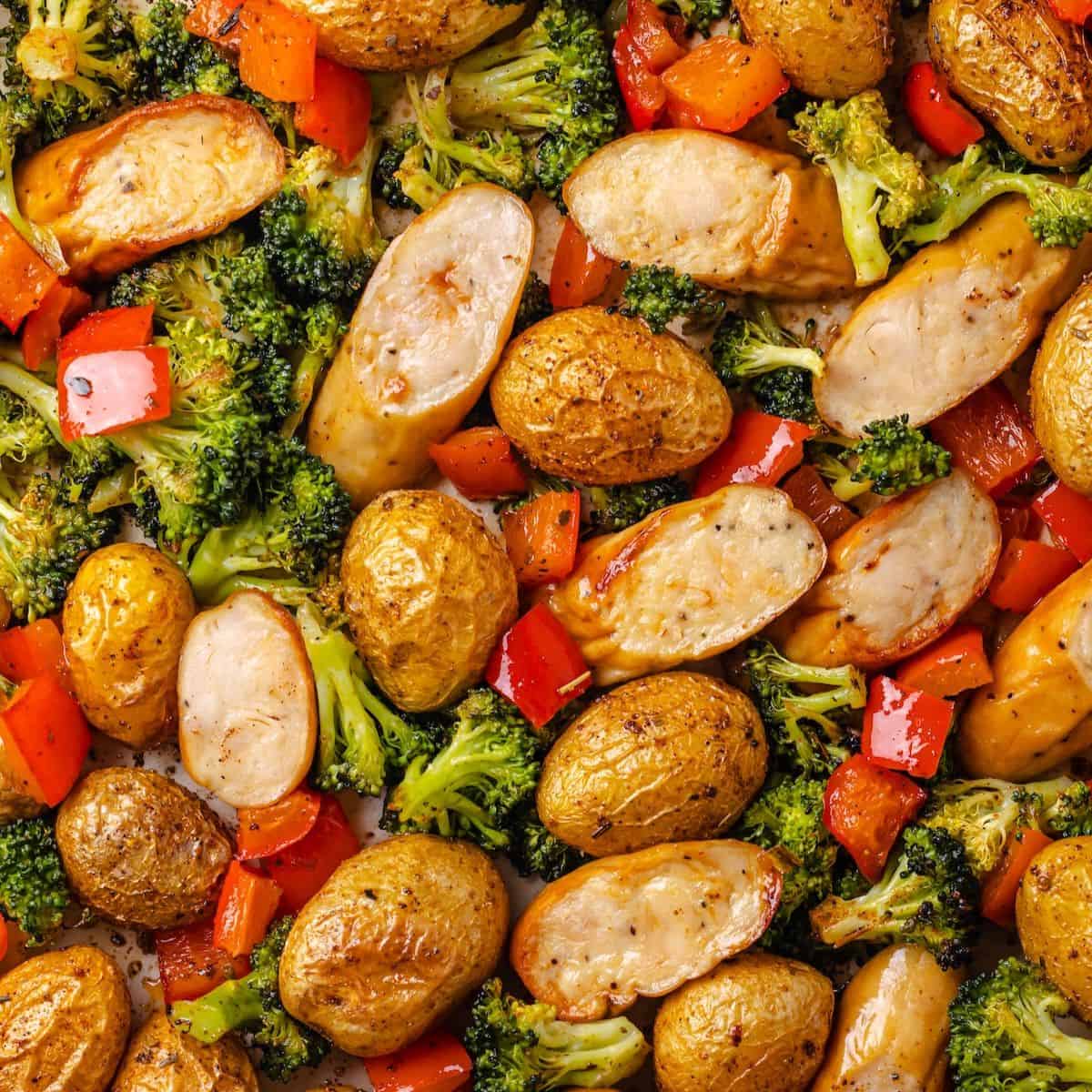 Close up of a sheet pan filled with chicken sausages, baby potatoes, red bell pepper and broccoli florets.