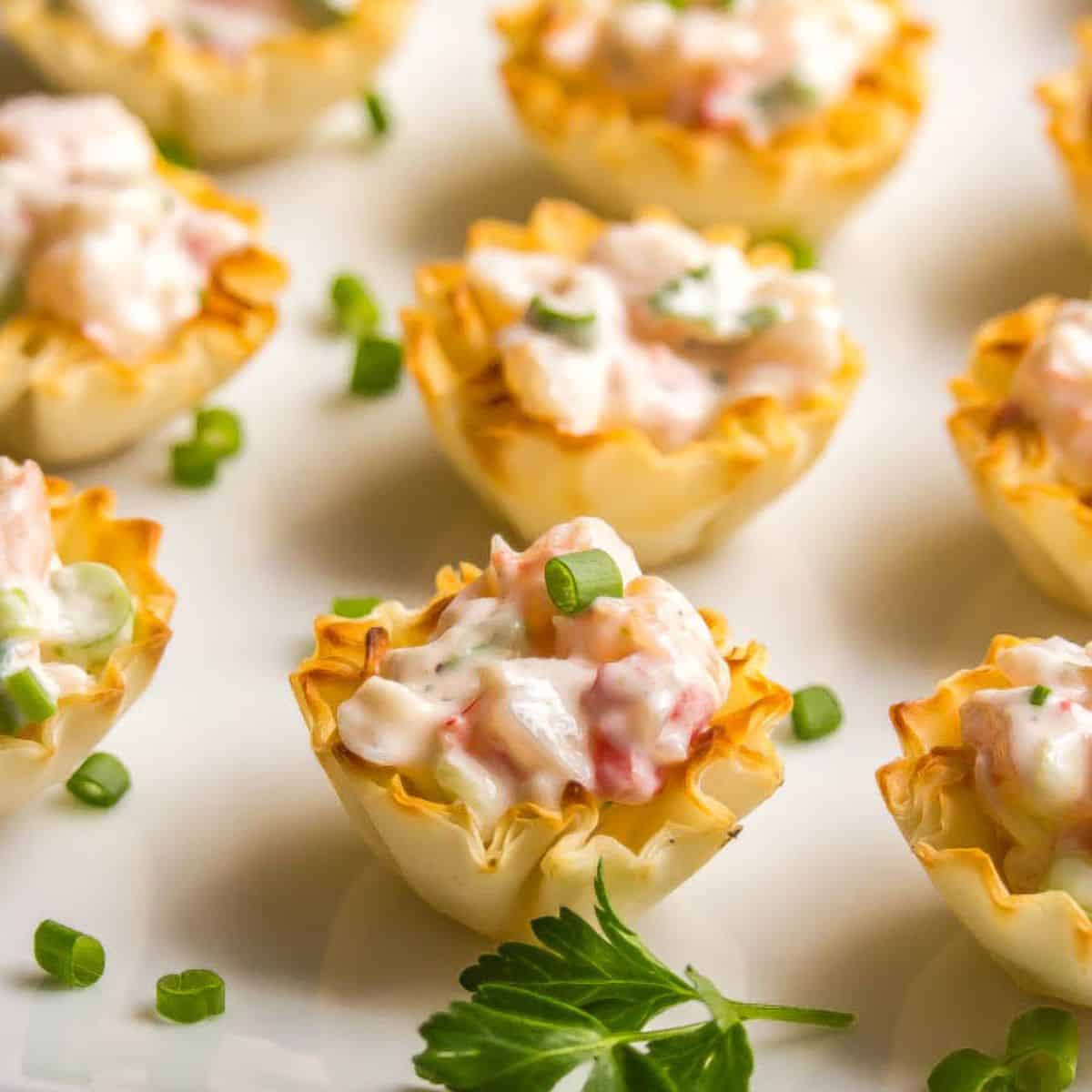Close up of mini phyllo cups filled with a creamy shrimp salad mixture and served on a white plate iwth parsley and sliced green onions for garnish.