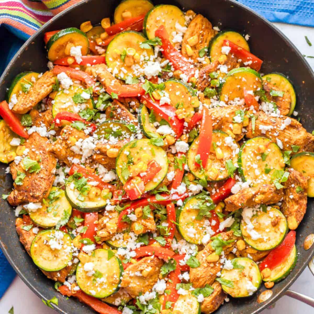 Close up of a skillet full of chicken strips and sliced zucchini with red peppers and corn and cotija cheese sprinkled on top.