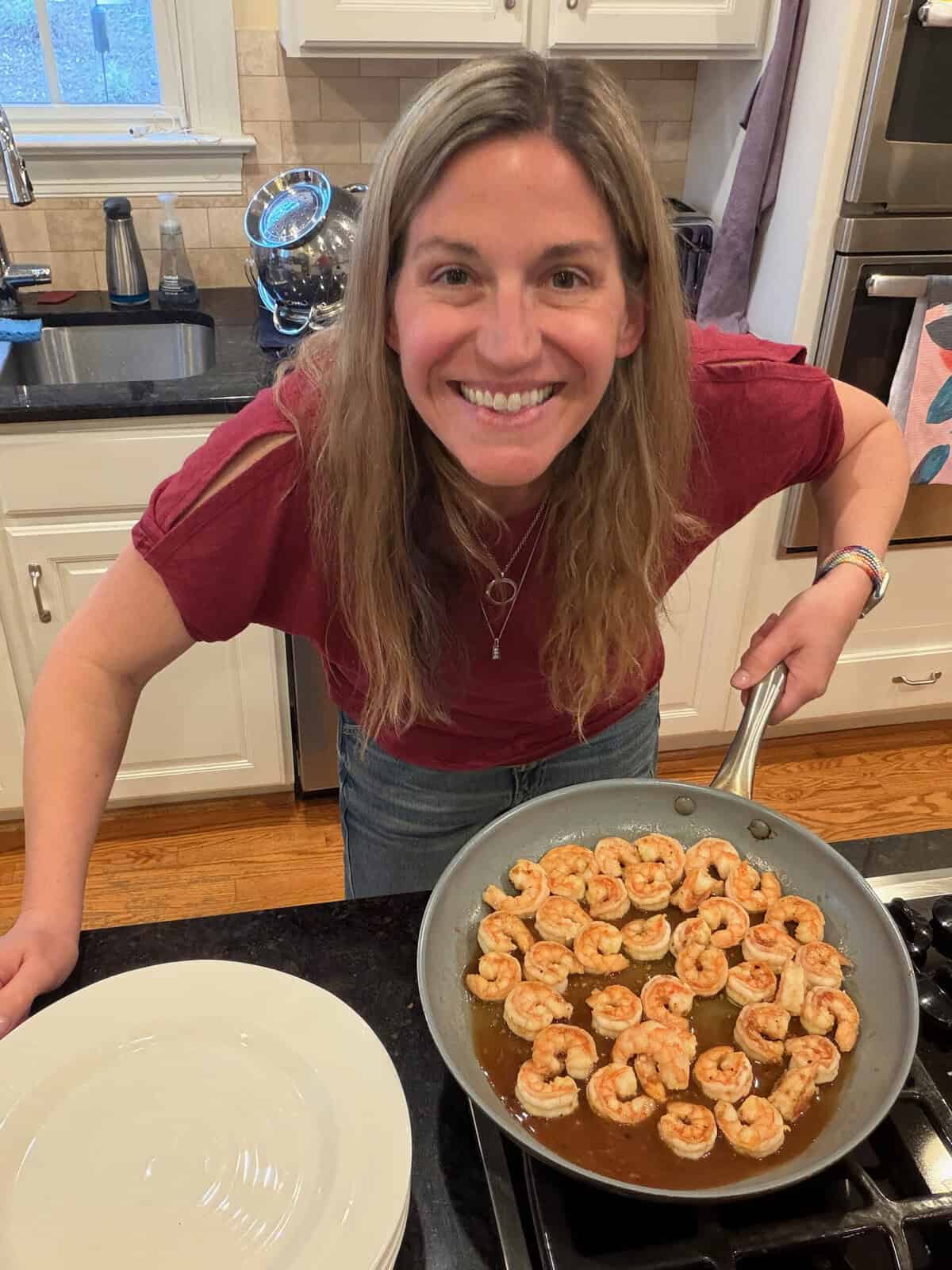 A woman in a red shirt holding up a skillet with saucy, cooked honey garlic shrimp with plates nearby for serving.