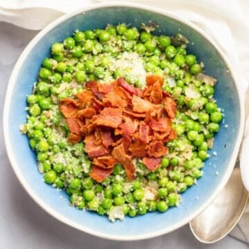 A blue tinted bowl filled with peas mixed with shallots and Parmesan cheese adn topped with cooked, crumbled bacon.