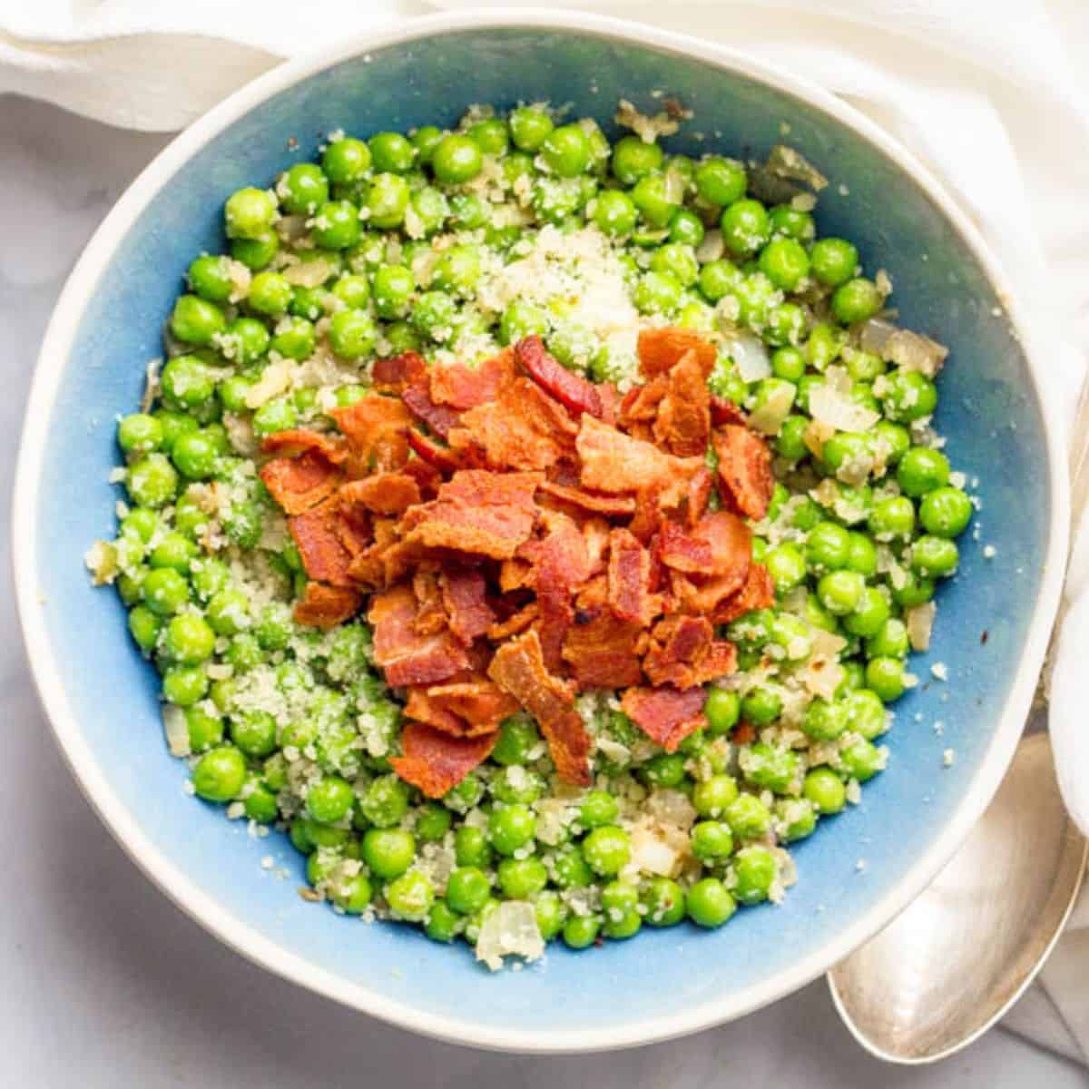 A blue tinted bowl filled with peas mixed with shallots and Parmesan cheese adn topped with cooked, crumbled bacon.