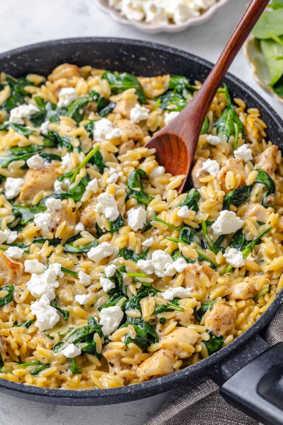 A wooden spoon resting in a dark skillet full of a creamy chicken, orzo and spinach mixture with feta cheese on top.