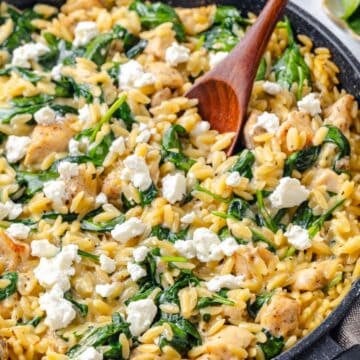 Close up of a wooden spoon resting in a dark skillet full of a creamy chicken, orzo and spinach mixture with feta cheese on top.