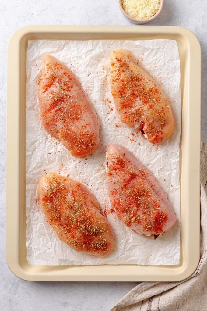 Cream cheese stuffed chicken breasts on a parchment paper lined baking sheet with seasonings on top.