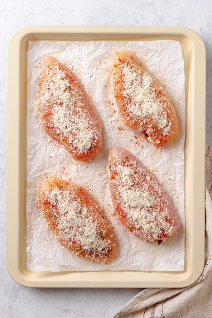 Cream cheese stuffed chicken breasts on a parchment paper lined baking sheet with seasonings and Parmesan cheese on top.