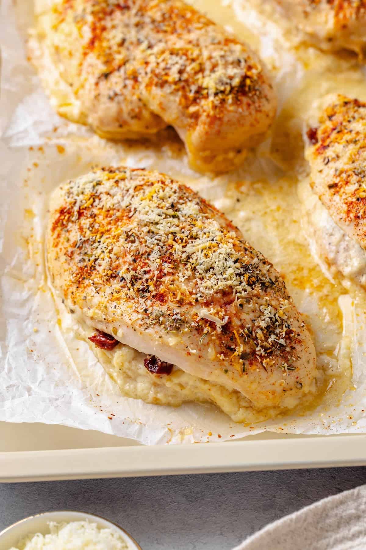 Chicken breasts stuffed with cream cheese and sun dried tomatoes with seasonings and Parmesan cheese on top on a baking sheet.