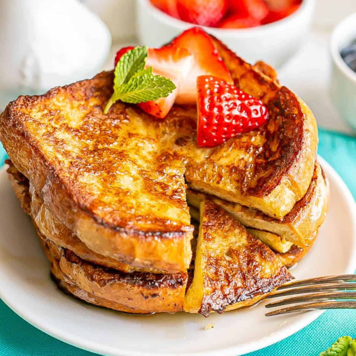 Close up of a stack of French toast on a small white plate with strawberries on top and a triangle bite section cut out.