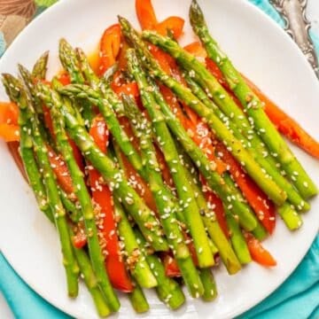 Close up of a white round plate with a mixture of asparagus and red pepper strips with sesame seeds sprinkled on top.