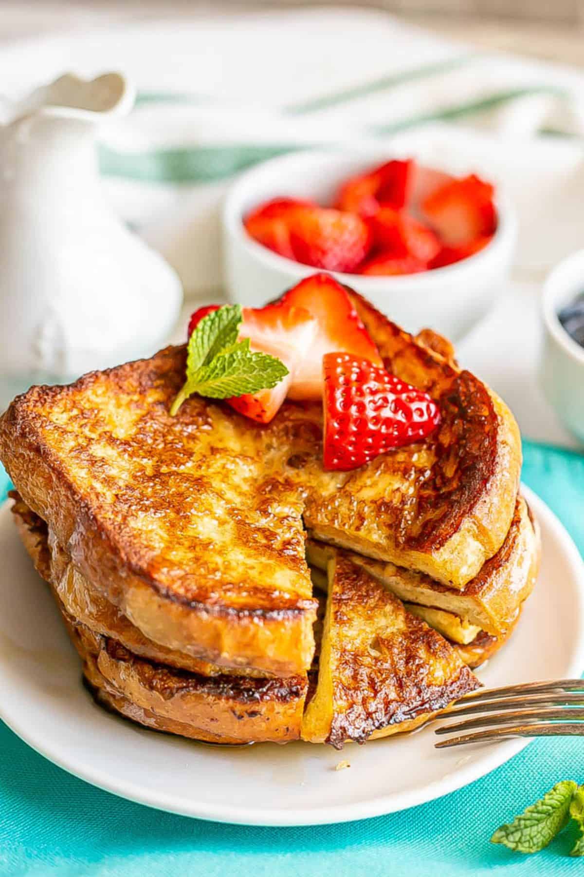 A stack of French toast on a small white plate with strawberries on top and a triangle bite section cut out.
