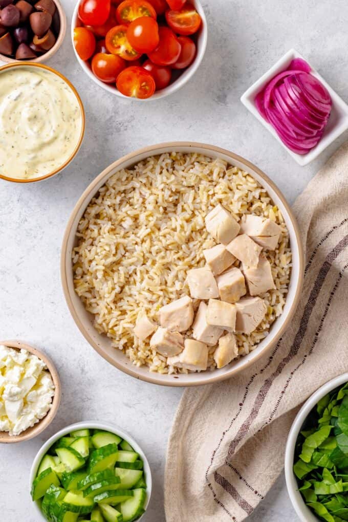 A bowl filled with brown rice with a pile of rotisserie chicken on one side and other veggies and toppings in bowls to the side before being added.