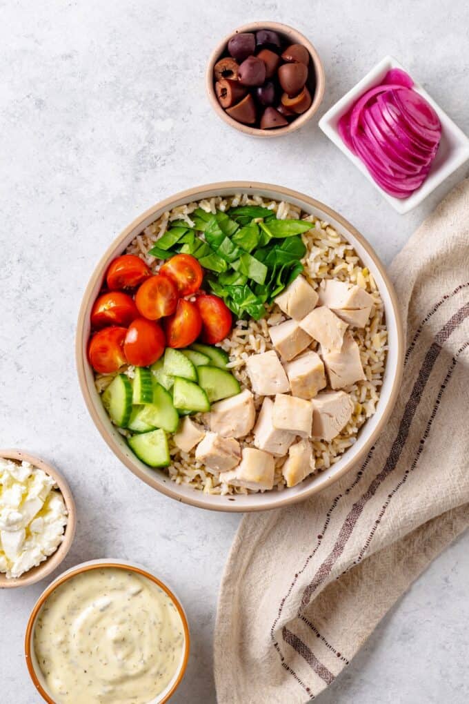 A bowl filled with brown rice with piles of rotisserie chicken, spinach, tomatoes and cucumber with additional toppings in bowls to the side before being added.