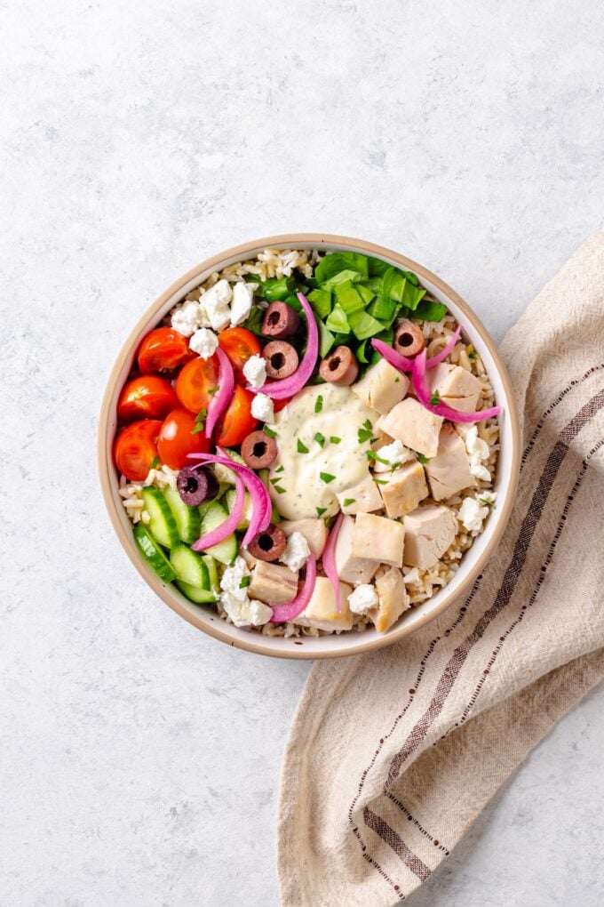 An assembled Greek chicken bowl with rice, spinach, tomatoes, cucumber, olives and tzatziki sauce in the middle.
