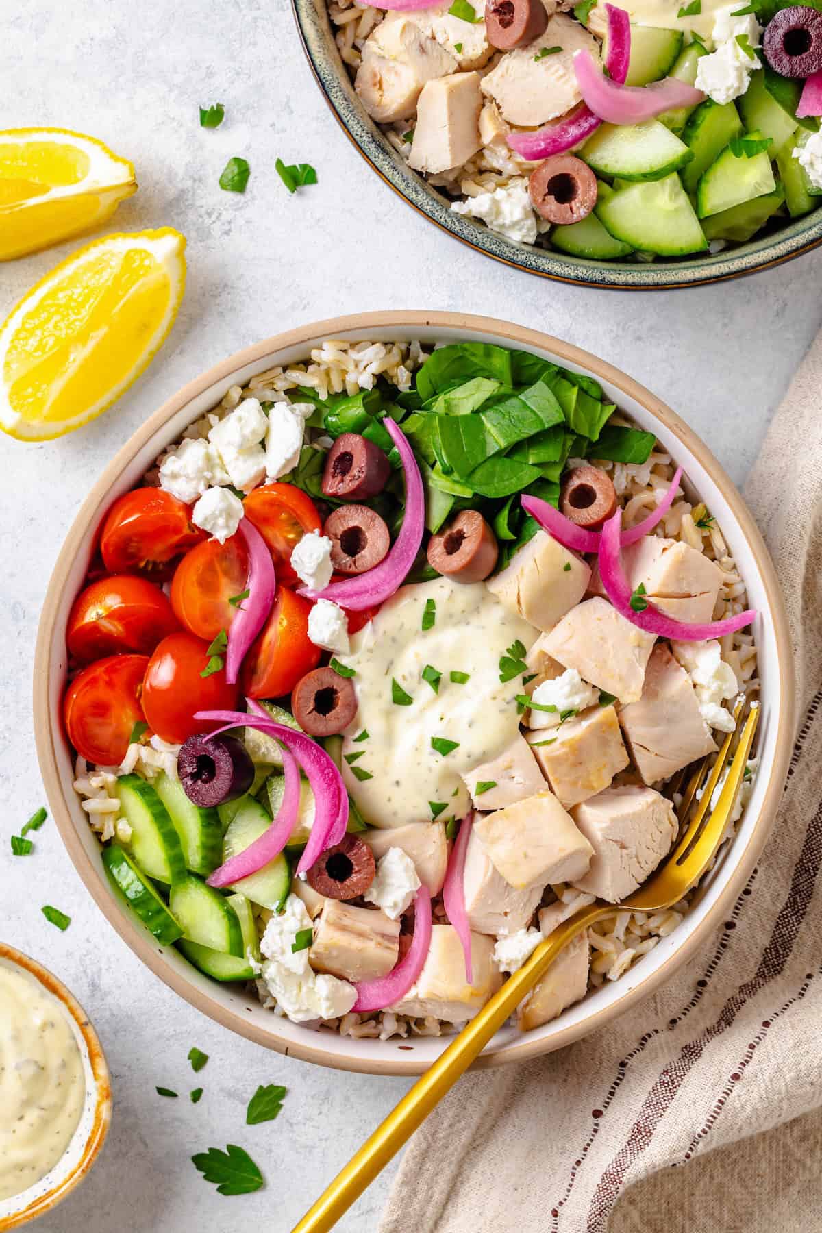 An arranged Greek chicken bowl with rice, spinach, tomatoes, cucumber, olives and tzatziki sauce in the middle and a fork resting on the side.