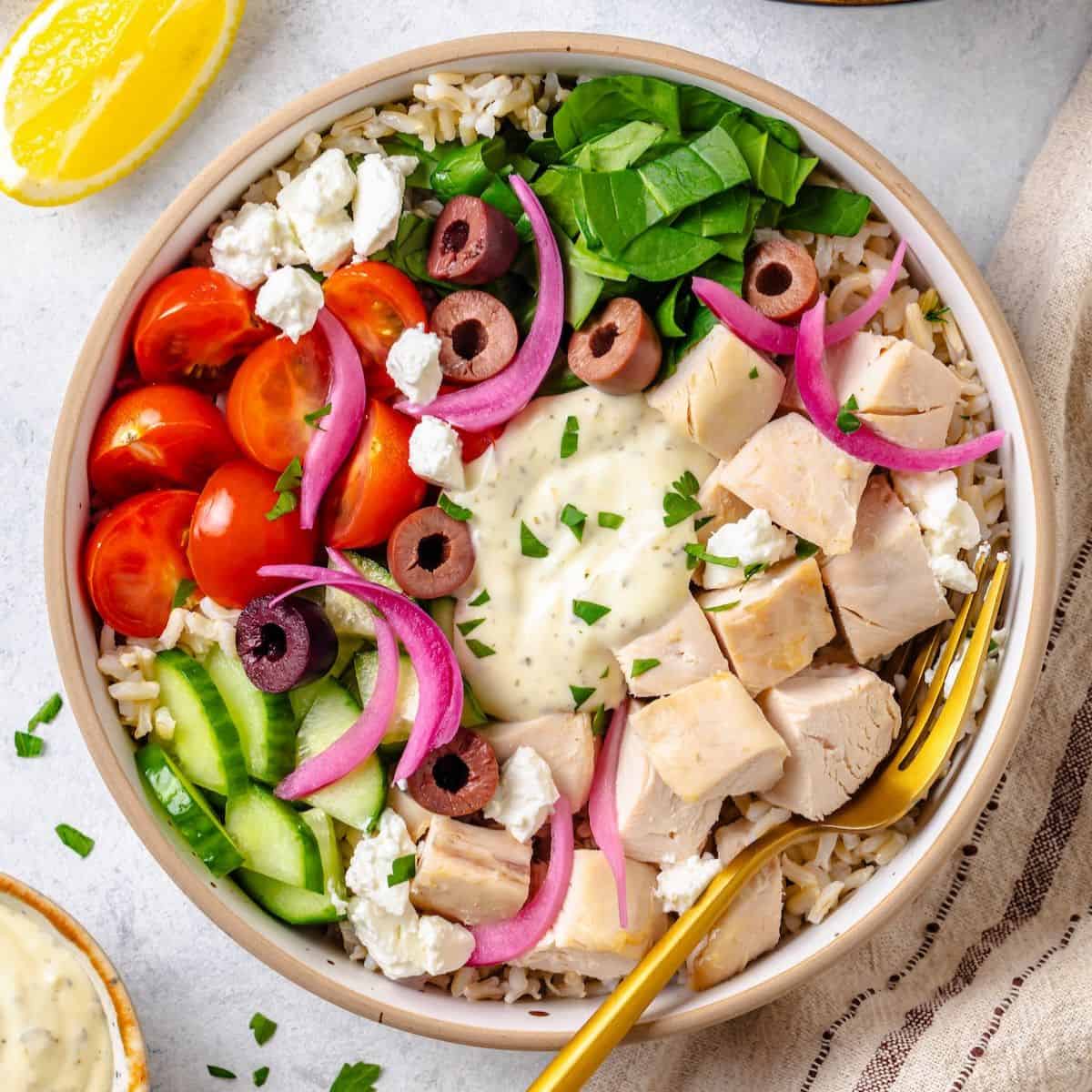 Close up of an arranged Greek chicken bowl with rice, spinach, tomatoes, cucumber, olives and tzatziki sauce in the middle and a fork resting on the side.