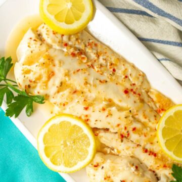 Close up of a long white platter with a piece of baked fish in Italian dressing with fresh parsley and lemon slices for garnish.