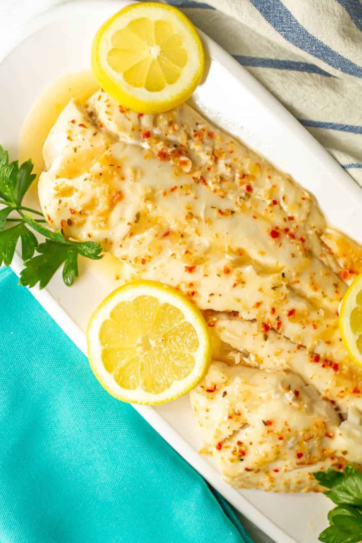 A long white platter with a piece of baked fish in Italian dressing with fresh parsley and lemon slices for garnish.