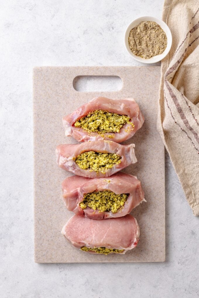 Four pork chops on a cutting board with a pesto and feta cheese mixture stuffed into pockets cut into each one.
