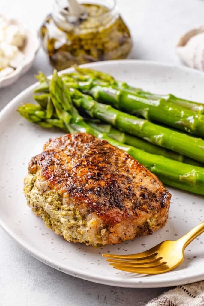 A pesto stuffed pork chop on a dinner plate with a fork to the side and steamed asparagus in the background.