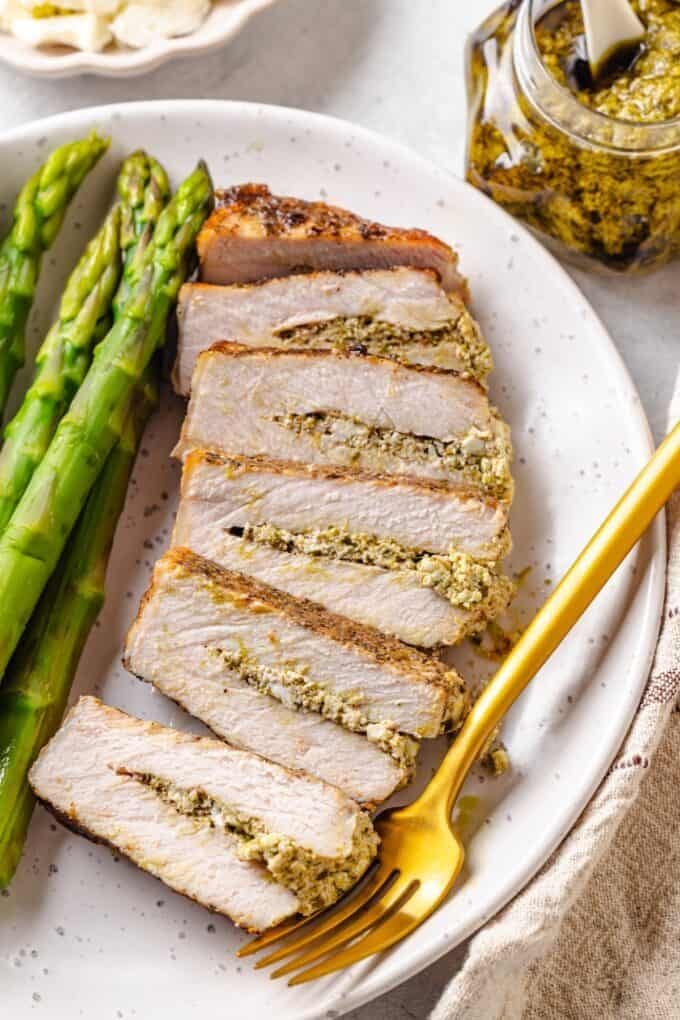 A sliced pesto stuffed pork chop on a dinner plate with a fork to the side and steamed asparagus alongside.