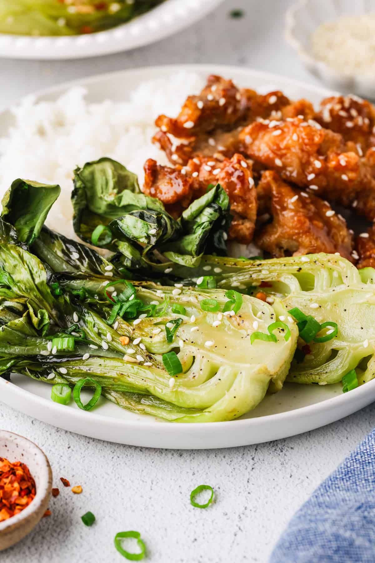 Two halves of roasted bok choy served alongside steamed white rice and a sticky chicken on a dinner plate.