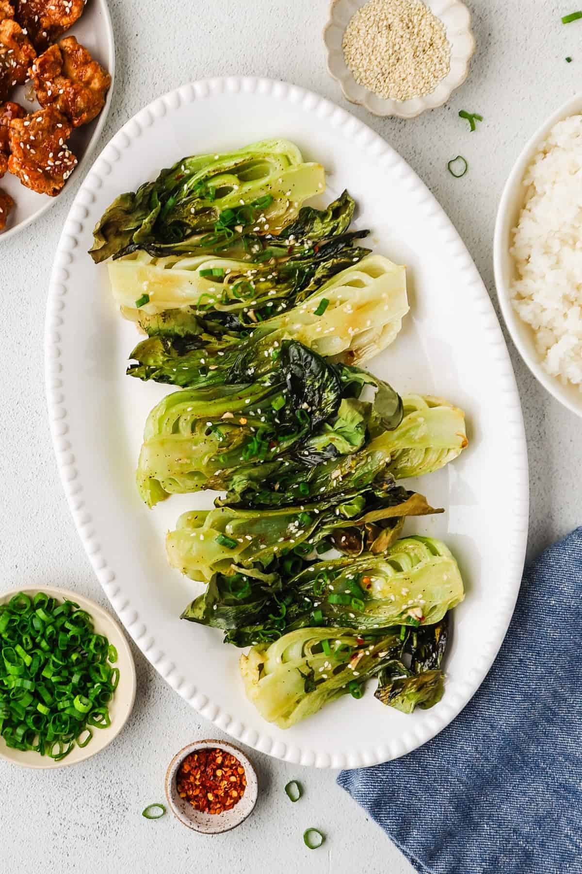 Roasted baby bok choy served on a white platter with bowls of sesame seeds, green onions and red pepper flakes nearby for topping, as well as bowls of rice and chicken for completing the meal.