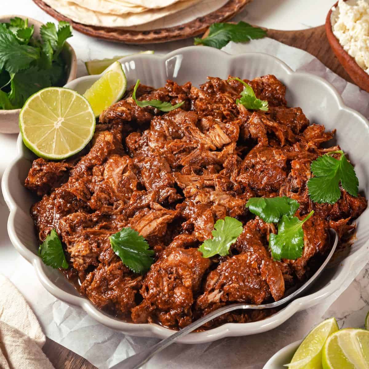 Close up of saucy mole chicken in a low serving bowl with cilantro and lime on top and a serving spoon to the side.