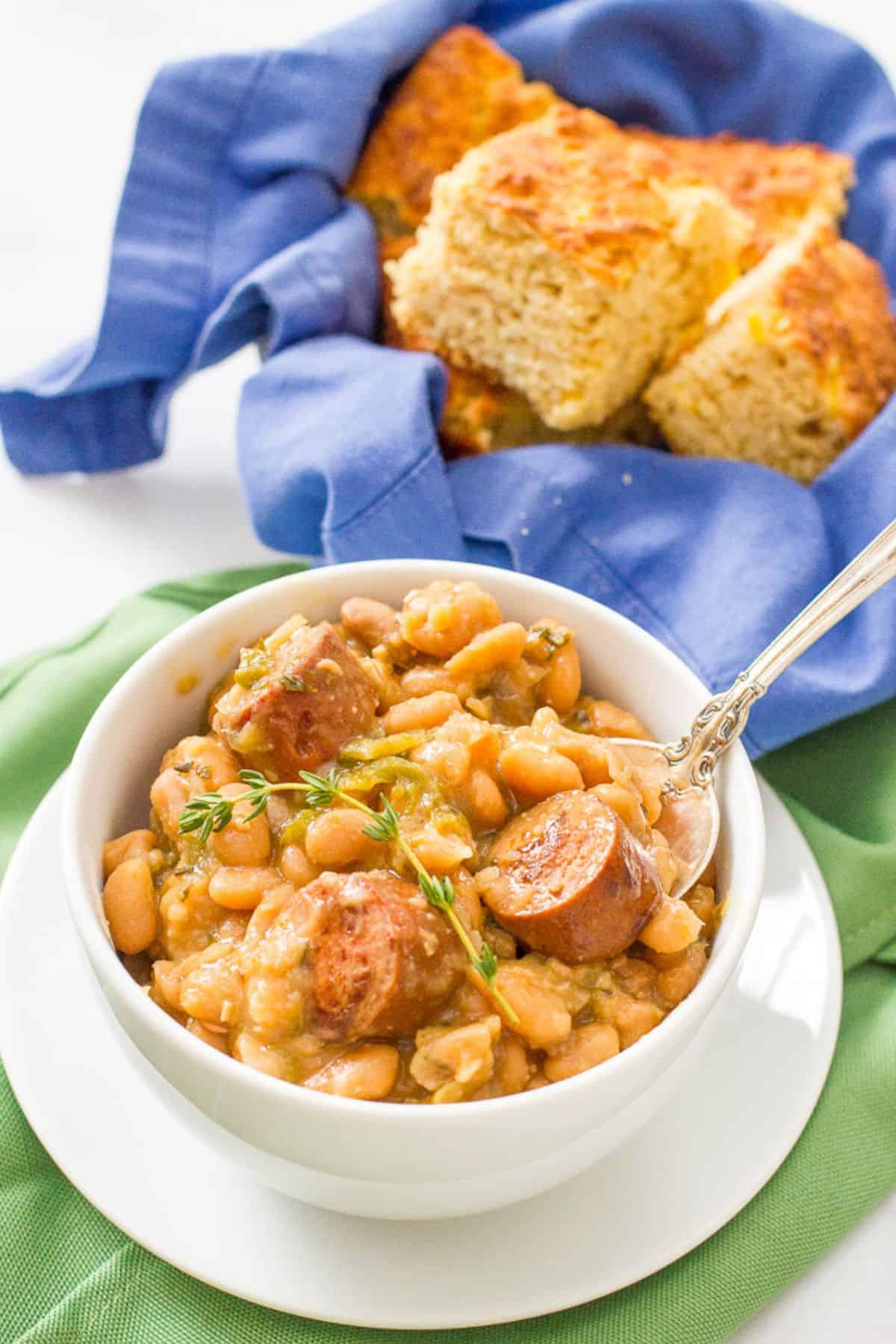 A white plate topped with a white bowl filled with creamy white beans and sliced sausage with a spoon resting in it and a bowl of cornbread in the background.