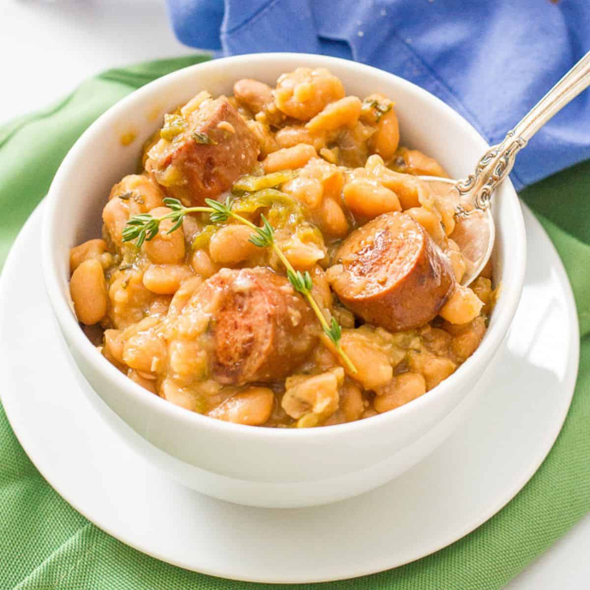 Close up of a white plate topped with a white bowl filled with creamy white beans and sliced sausage with a spoon resting in it.