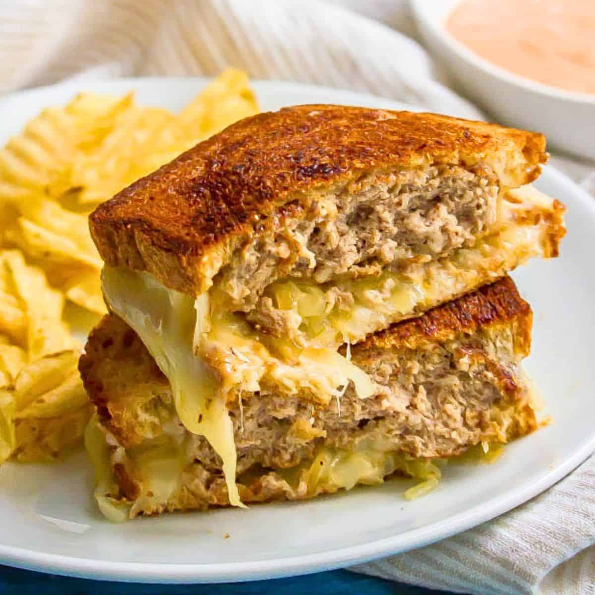 Close up of a stacked ground turkey patty melt sandwich with melty cheese on a white plate alongside some potato chips.