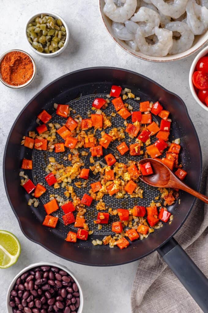 A large skillet with sautéed onion and red bell pepper with a wooden spoon resting in it.