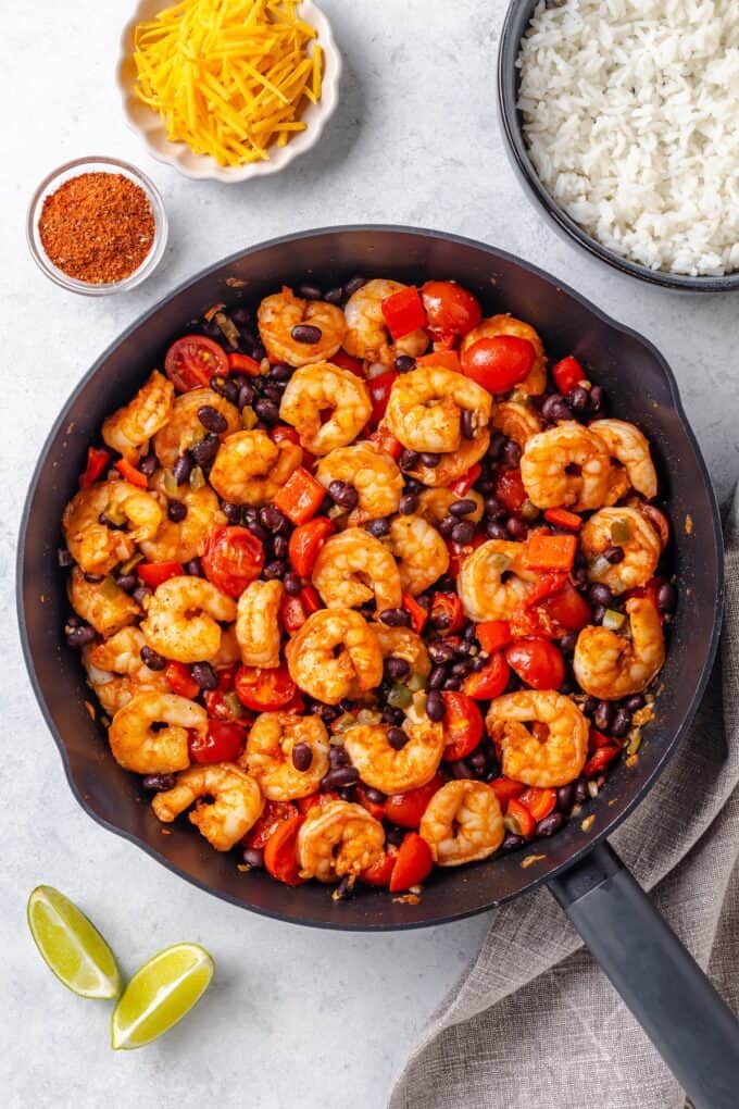 A large skillet with shrimp mixed with black beans, tomatoes and peppers with toppings to the side.