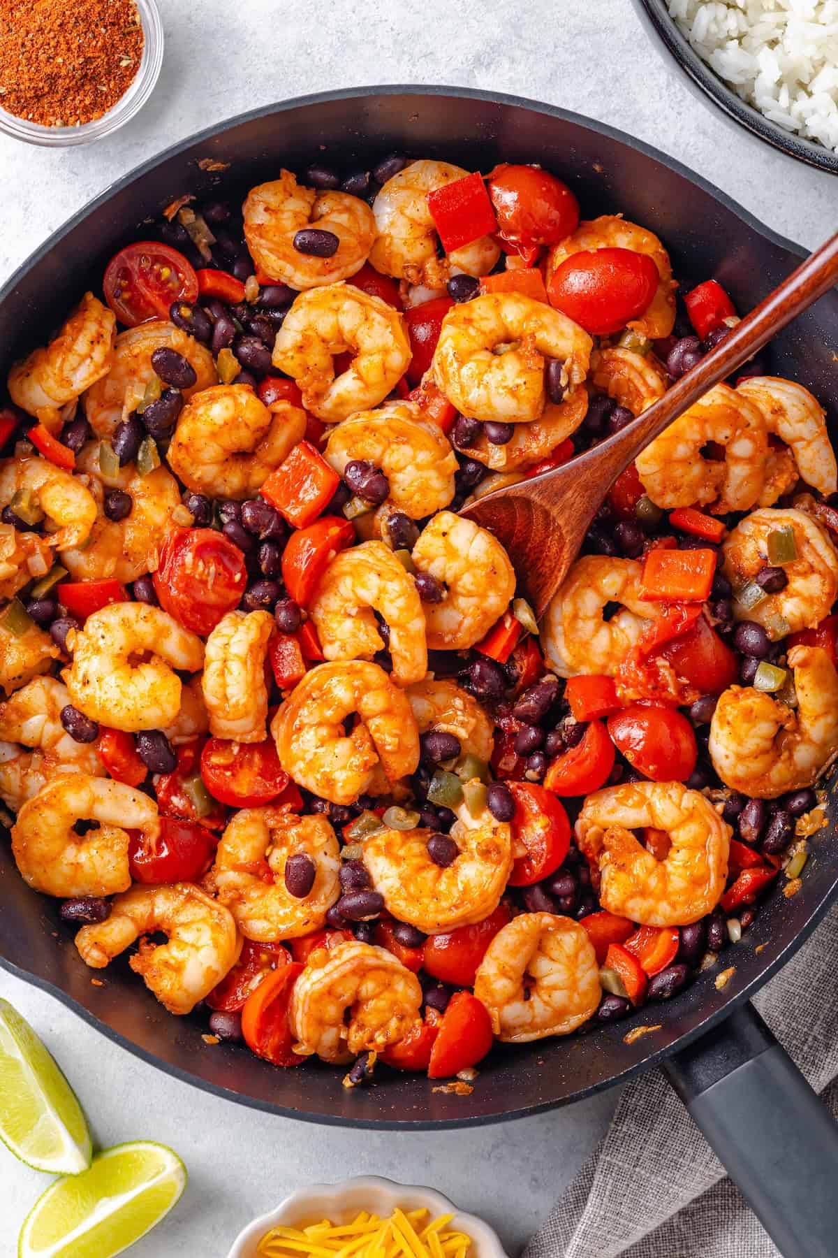 A wooden spoon resting in a large skillet with a mixture of shrimp, black beans, tomatoes and peppers.