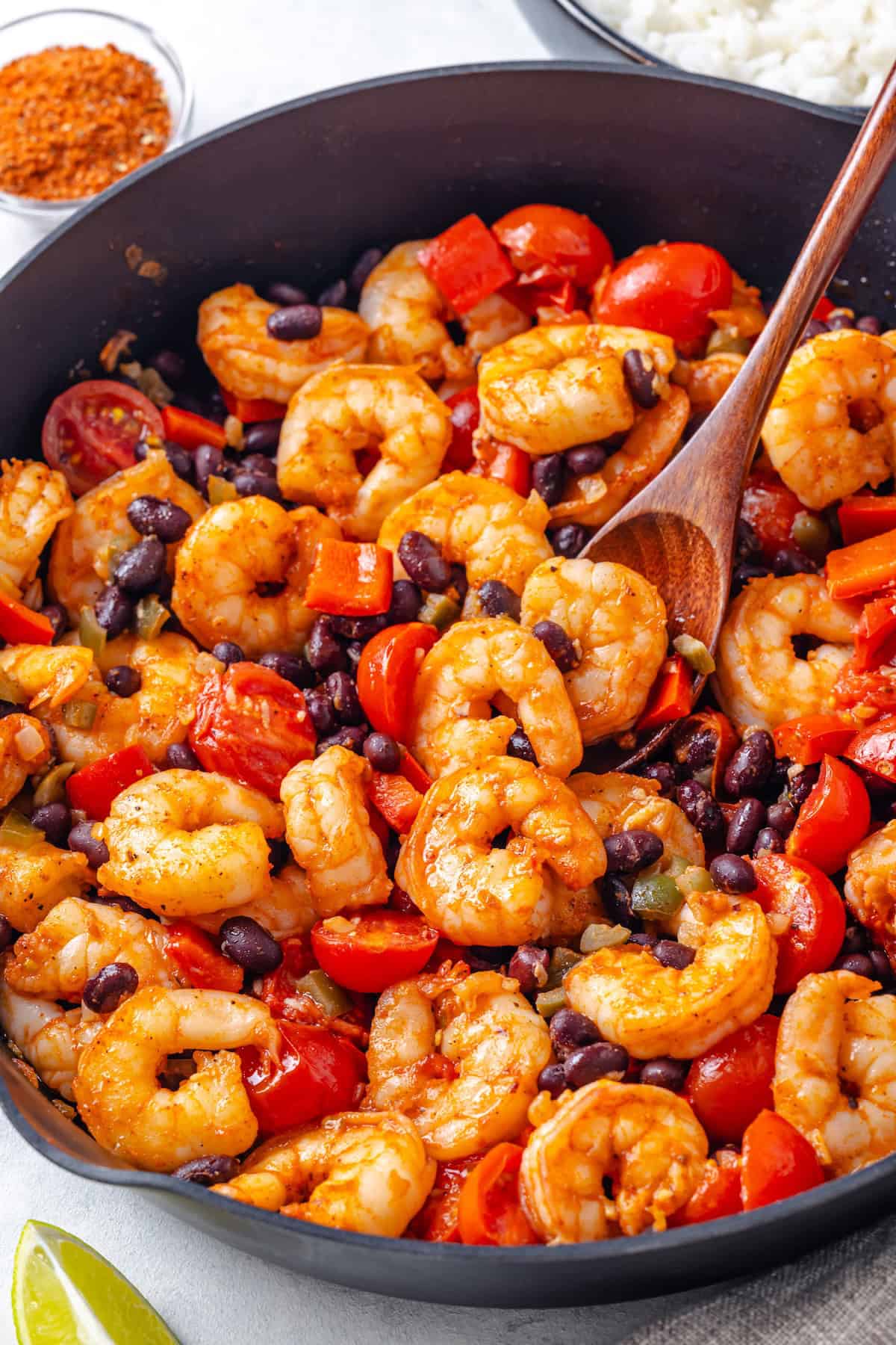 A wooden spoon resting in a large skillet with a mixture of shrimp, black beans, cherry tomatoes and peppers.