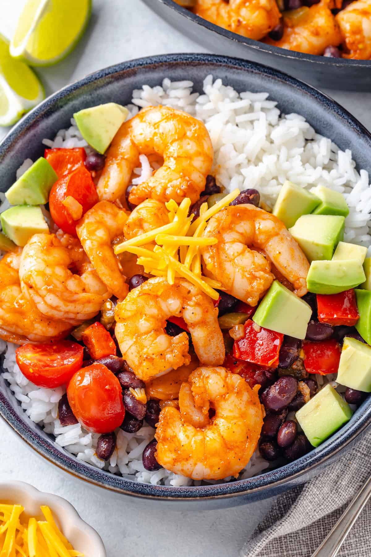 Southwest style shrimp with black beans and tomatoes served in a bowl over white rice with cheese and avocado for toppings.