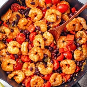 Close up of a wooden spoon resting in a large skillet with a mixture of shrimp, black beans, tomatoes and peppers.