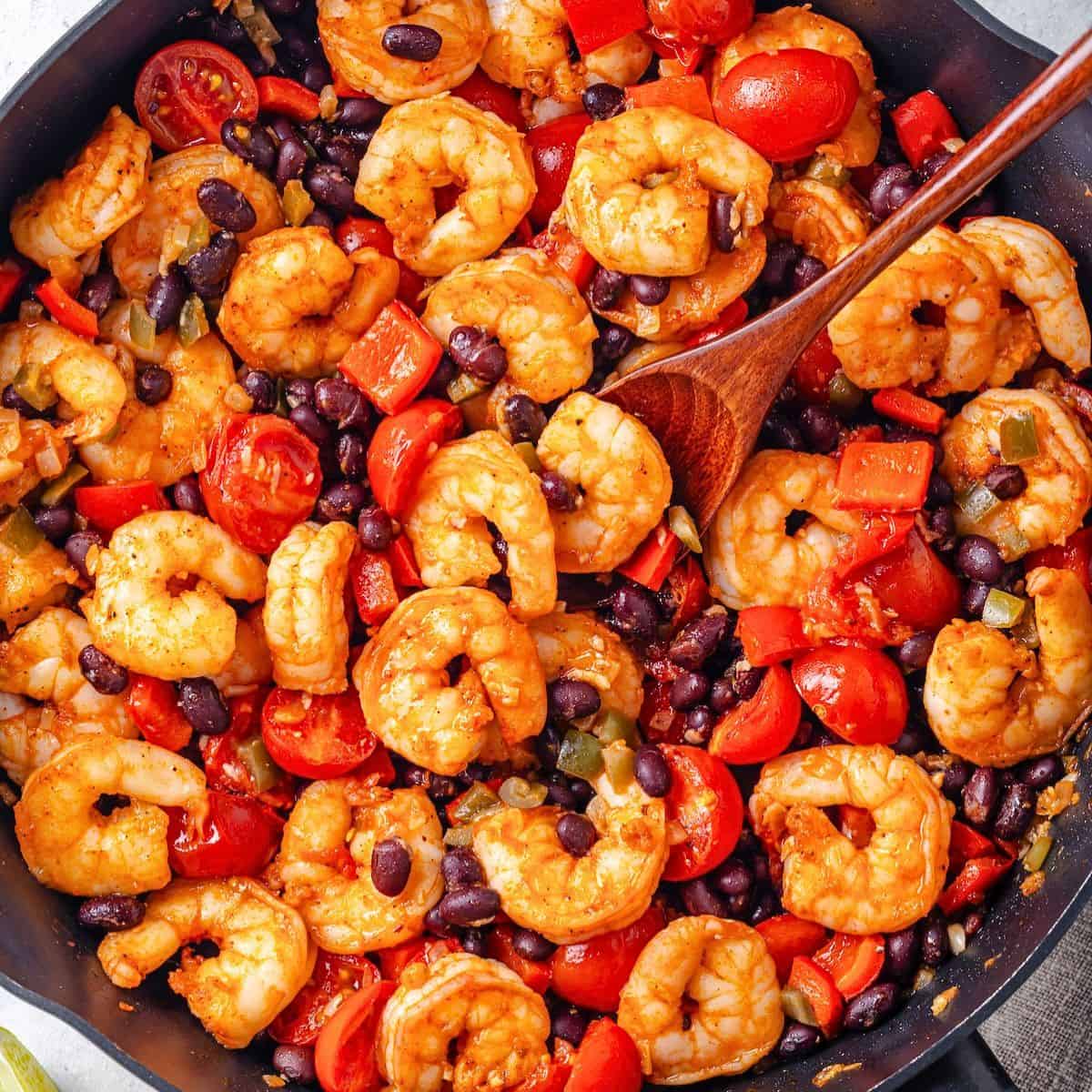 Close up of a wooden spoon resting in a large skillet with a mixture of shrimp, black beans, tomatoes and peppers.