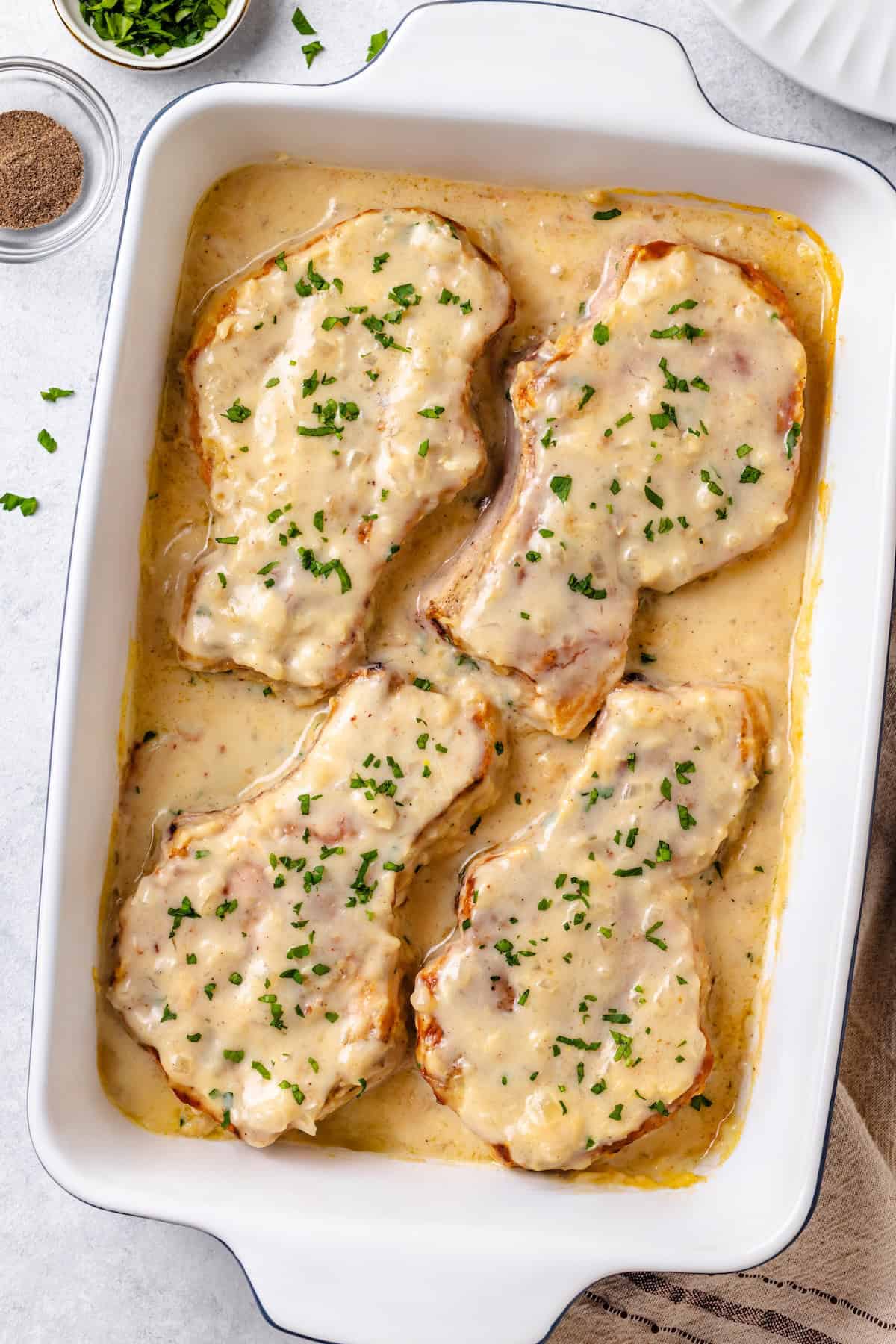 Baked smothered pork chops in a white baking dish with fresh parsley sprinkled on top.