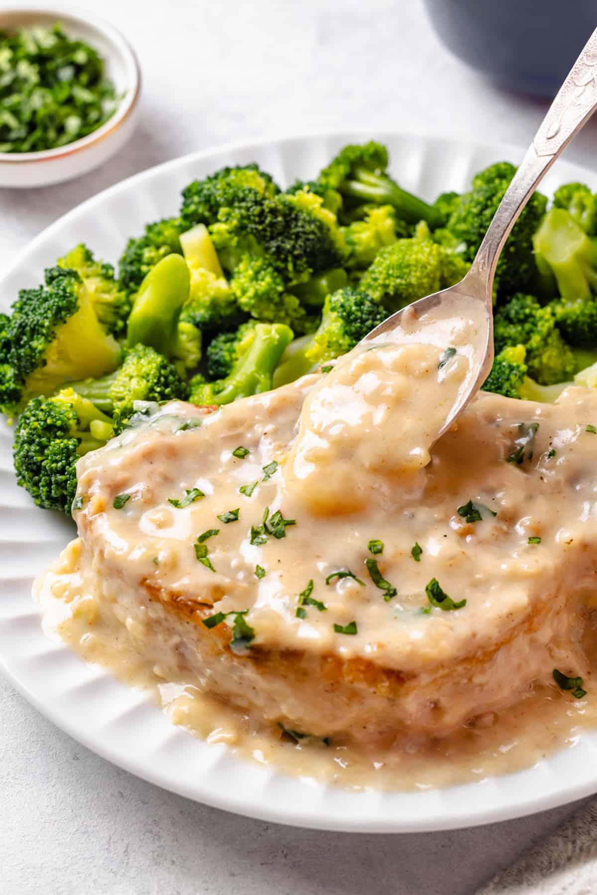 Gravy being spooned over a baked pork chop on a dinner plate alongside steamed broccoli.