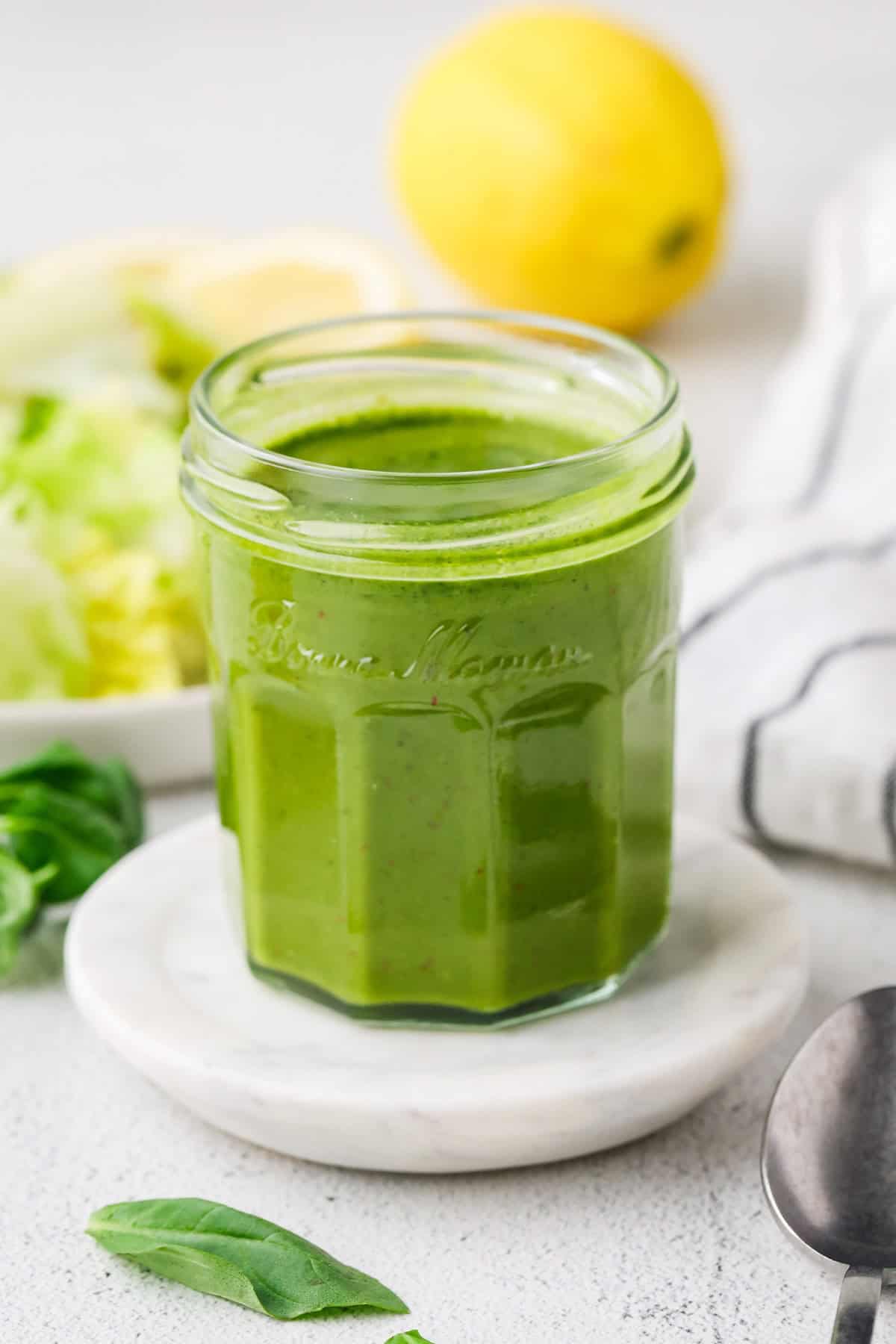 Fresh homemade basil vinaigrette in a glass jar set on a small stone with a lemon and salad in the background.