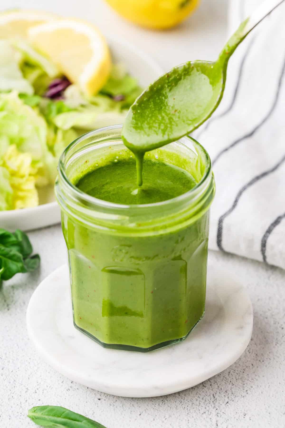 A spoon drizzling fresh homemade basil vinaigrette in a glass jar with a lemon and salad in the background.
